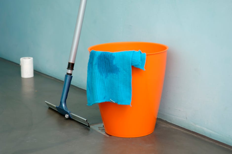 An indoor scene featuring a bright orange plastic bucket with a blue microfiber cleaning cloth draped over its rim, placed on a smooth, grey floor next to a floor squeegee with a long handle and rubbe